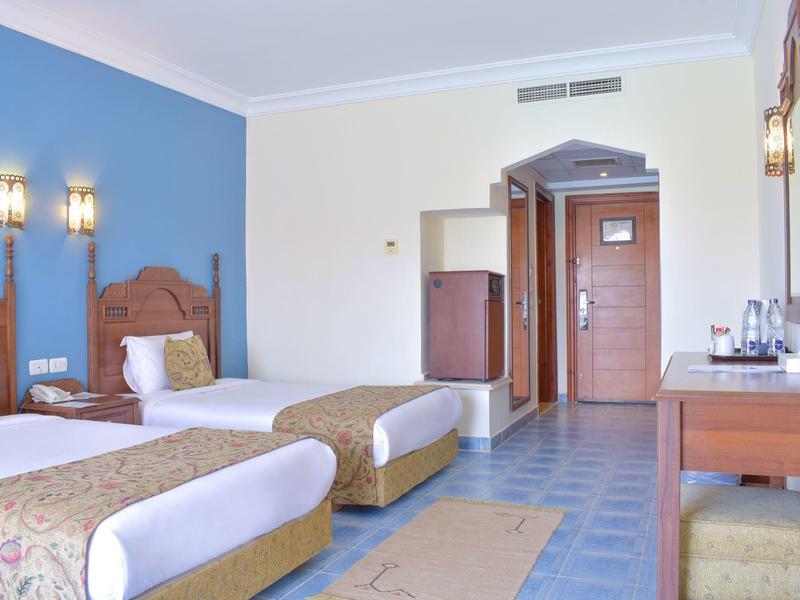 Zwei Einzelbetten mit Holzrahmen, blaue Wand, Schreibtisch und Eingangstür in hellem Hotelzimmer.