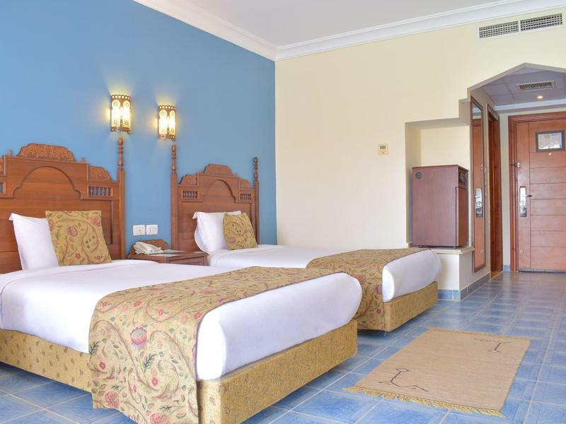 Hotelzimmer mit zwei Einzelbetten, blauer Wand, Holzmöbeln und hellblauem Fliesenboden.