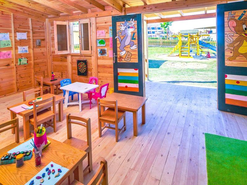 Helles Holz-Klassenzimmer mit kleinen Tischen, bunten Stühlen und offener Tür zum Spielplatz draußen.