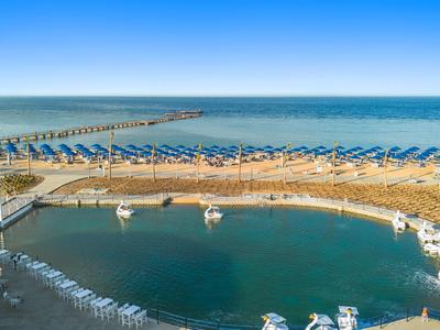 Strand mit gelben Sonnenschirmen, blauem Meer und einem kleinen Hafen mit Booten im Vordergrund.