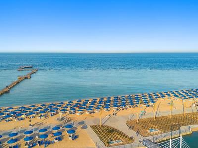 Strand mit vielen blauen Sonnenschirmen und steigendem Holzsteg ins Meer unter klarem Himmel.