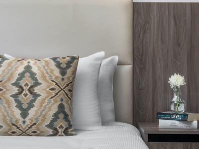 Bett mit gemustertem Kissen, weißen Kissen und Nachttisch mit Vase, Buch und Lampe.