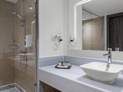 Hell beleuchtetes Badezimmer mit rechteckigem Spiegel, weißem Waschbecken und Glasduschabtrennung.