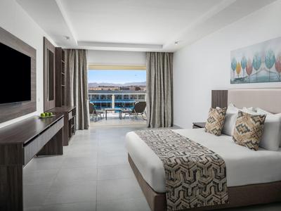 Modernes Hotelzimmer mit großem Bett, TV, Kommode und Blick auf Terrasse und Meer durch bodentiefe Fenster.