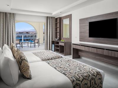 Helle Hotelzimmer mit zwei Betten, Fernseher, Schreibtisch und Balkon mit Meerblick.