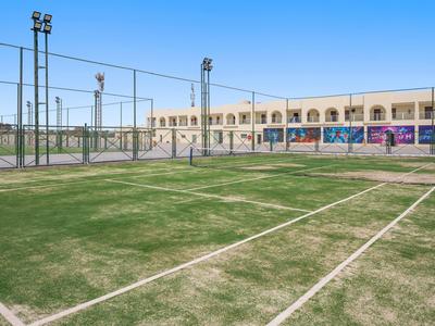 Freiluft-Tennisplatz mit grünem Kunstrasen, umgeben von Zäunen und Schulgebäuden im Hintergrund.