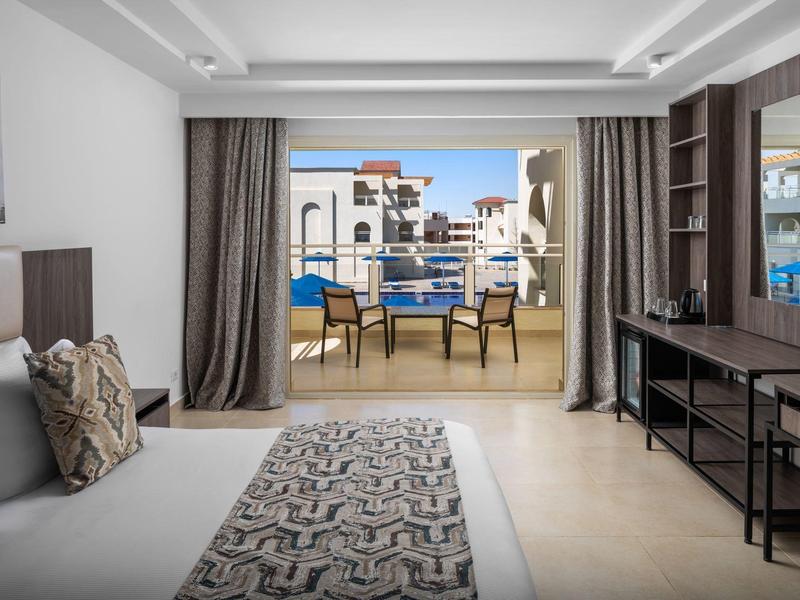 Modernes Hotelzimmer mit Bett, Schreibtisch, Balkon mit Stühlen und Blick auf Gebäude.