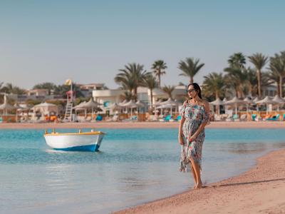 Frau in Sommerkleid läuft barfuß am Strand mit Boot und Palmen im Hintergrund.