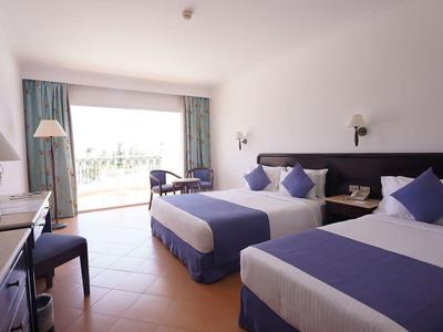 Bright hotel room with two beds, desk, chair, and view of a terrace.