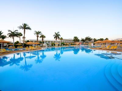 Groot zwembad met palmbomen en ligstoelen bij een hotelresort onder een heldere hemel.