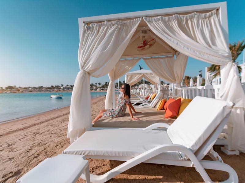 Beach with white lounge chairs and canopy beds on sandy shore under clear sky.