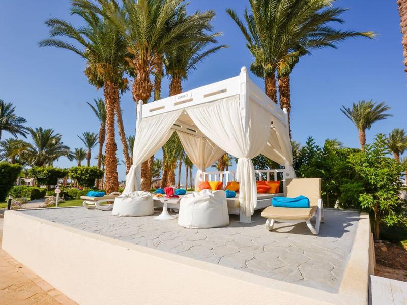White lounge with curtains and cushions under palm trees on a clear sky day.