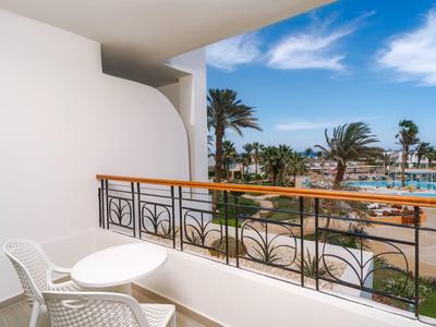 Balkon z dwoma białymi krzesłami i małym stolikiem z widokiem na basen i palmy pod niebieskim niebem.