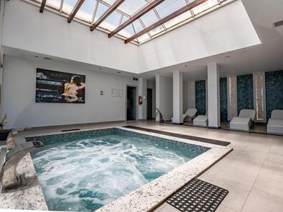 Basen kryty z jacuzzi, białymi leżakami i szklanym dachem wpuszczającym naturalne światło.