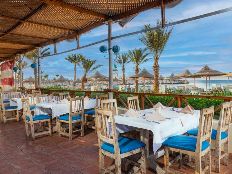 Otwarta przestrzeń restauracyjna z białymi stołami i niebieskimi krzesłami pod dachem, widok na morze.