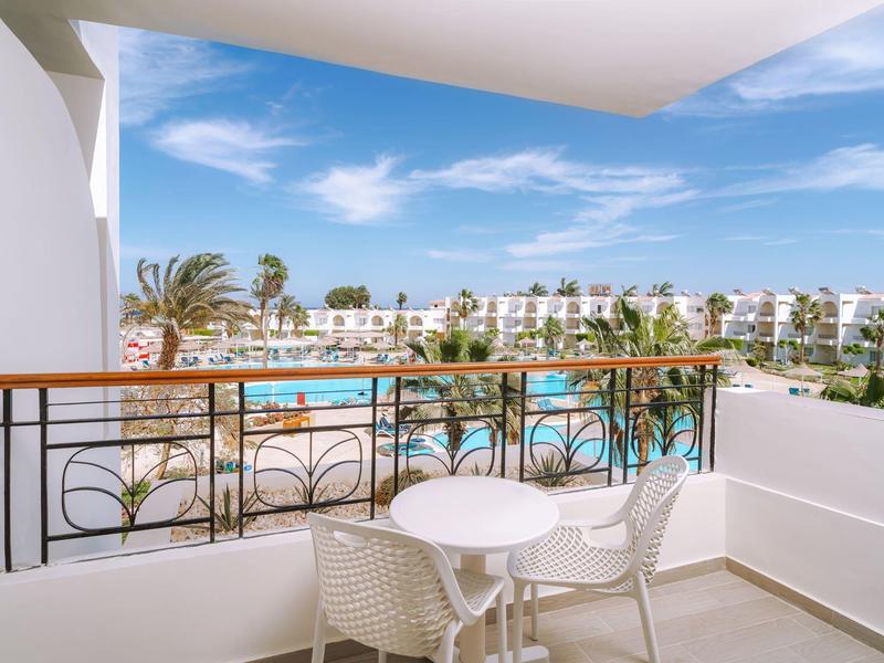 Balkon z dwoma krzesłami i stołem z widokiem na basen hotelowy i białe budynki pod niebieskim niebem.