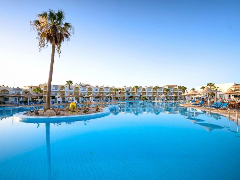 Duży hotelowy basen z wyspą palmową i leżakami pod bezchmurnym niebem.