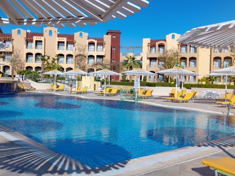 Duży basen hotelowy z leżakami i parasolami przed wielopiętrowymi budynkami pod czystym niebem.