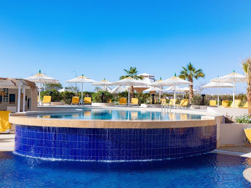 Okrągły basen z niebieskimi kafelkami i parasolami w zewnętrznej części hotelu.