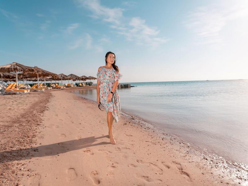 Kobieta w letniej sukience spacerująca po piaszczystej plaży z parasolami i spokojnym morzem o wschodzie słońca.