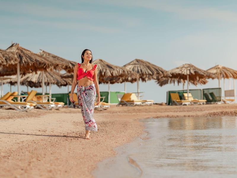 Kobieta w czerwonym topie spaceruje po plaży z parasolami z trzciny i leżakami w tle.