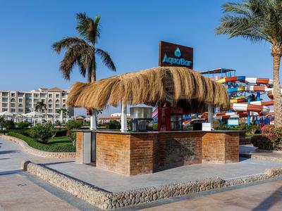 Een kleine strandbar met rieten dak en een palmboom, omgeven door hotels en glijbanen.