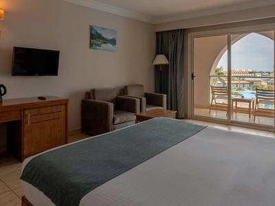 Bright hotel room with double bed, seating area, and balcony overlooking the harbor.