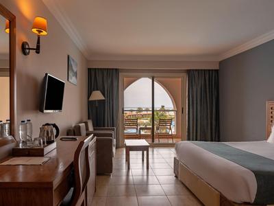 Bright hotel room with double bed, desk, chairs, and balcony view overlooking the sea.