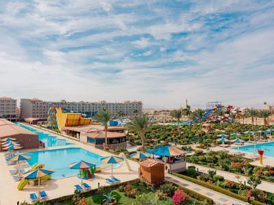 Large resort with multiple pools, sun umbrellas, and surrounding hotels under a cloudy sky.