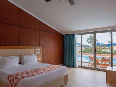 Modern hotel room with large bed and view of pool and palm trees through glass windows.
