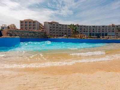 Kompleks hotelowy z dużym basenem i piaszczystą plażą pod zachmurzonym niebem.
