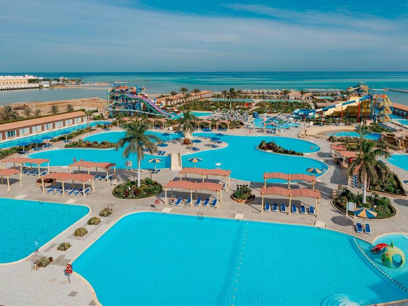 Duży kompleks hotelowy z wieloma basenami, palmami, leżakami i widokiem na morze przy czystym niebie.