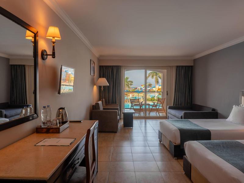 Chambre d'hôtel avec deux lits, balcon avec vue sur les palmiers et la mer, éclairage chaleureux.