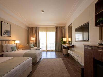 Modern hotel room with two beds, TV, desk, and balcony with daylight.