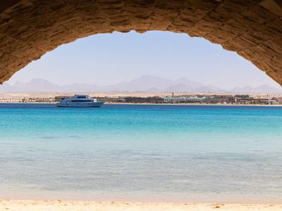 View through a stone arch at turquoise water with boat and distant mountains.