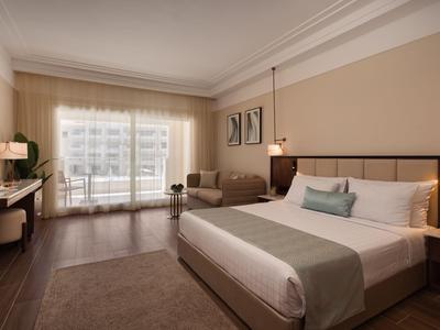 Modern hotel room with large bed, sofa, and balcony in daylight.