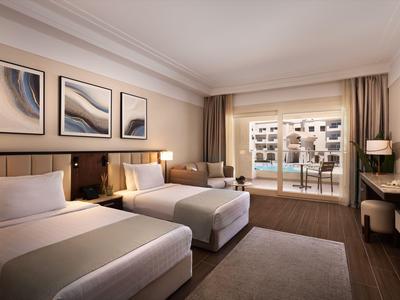 Modern hotel room with two beds, balcony, and view of surrounding buildings.