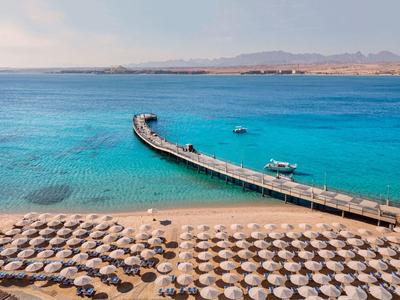 Long pier extends into turquoise water with sandy beach and many umbrellas.