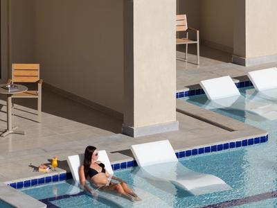 A person relaxes in a modern pool with loungers in the water next to light-colored walls.