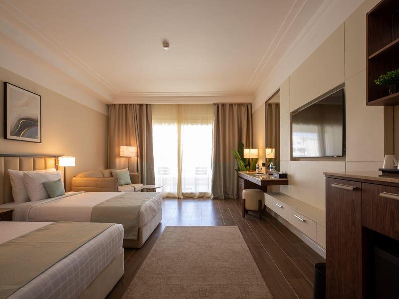 Modern hotel room with two beds, TV, desk, and balcony with daylight.