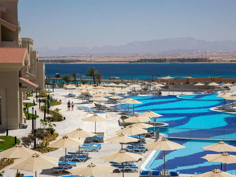 Large hotel pool area with loungers and umbrellas, sea and mountains in the background.