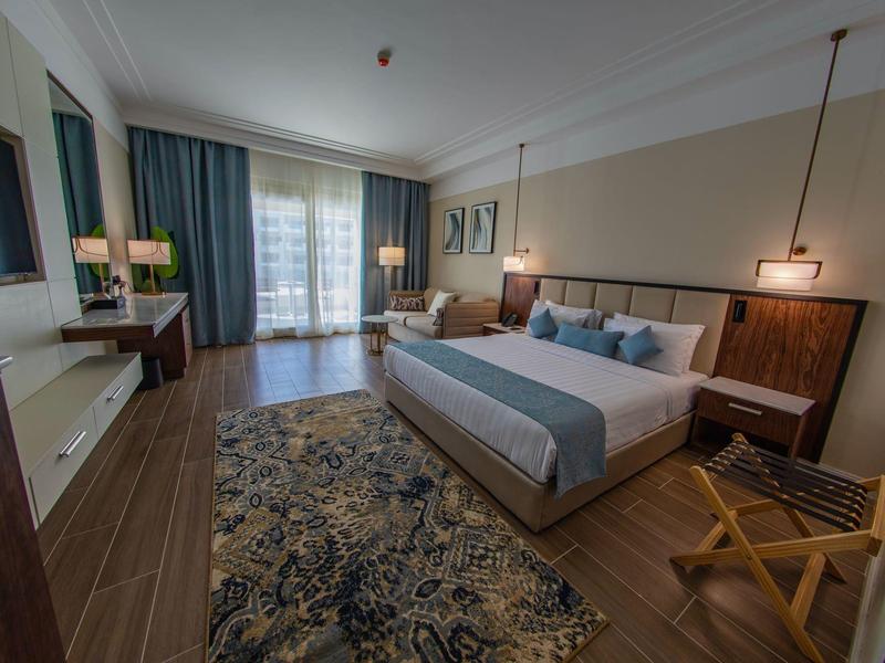 Modern hotel room with large bed, desk, chairs, and balcony with curtains.