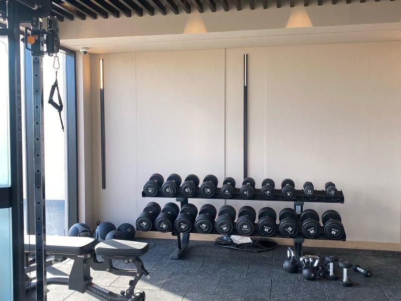 Moderner Fitnessraum mit Hanteln, Trainingsbank und Kraftgerät neben großen Fenstern.