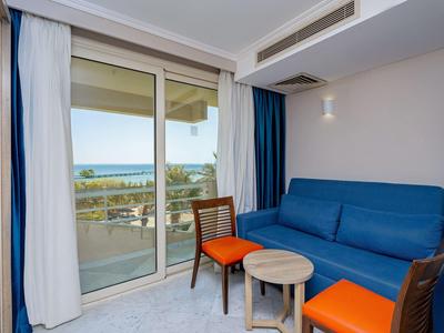 Chambre d'hôtel lumineuse avec canapé bleu, deux chaises aux coussins orange et vue sur la mer depuis le balcon.