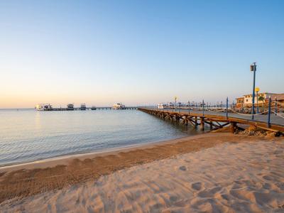 Une longue jetée en bois s'étend sur une eau calme au coucher du soleil, avec une plage de sable au premier plan.