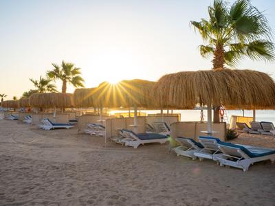 Sonnenuntergang am Strand mit Palmen und Liegestühlen unter Strohdächern.