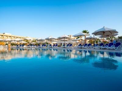 Großer Hotelpool mit blauen Liegestühlen und Sonnenschirmen im Sonnenlicht.