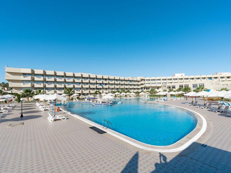Grand hôtel avec grande piscine extérieure et chaises longues sous un ciel bleu clair.
