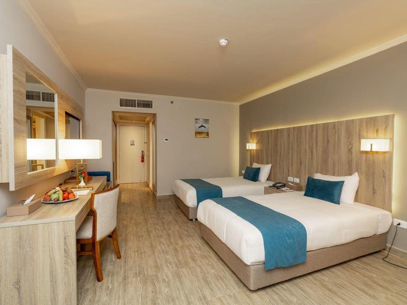 Chambre d'hôtel lumineuse avec deux lits simples, bureau et chaise, éclairage moderne et sol en bois.