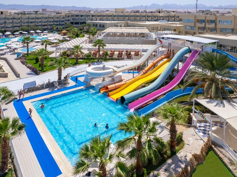 Grande piscine d'hôtel avec toboggans colorés, parasols et palmiers dans un cadre ensoleillé.